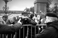 Black-and-white photo of a man in a flat cap and fingerless gloves leaning on a metal crowd barrier, hands cupped around his mouth as he shouts at a uniformed officer in a beret and face mask, seen from behind in the right foreground. A blurred arm with a pointing hand enters from the left. Behind the barrier, a crowd watches, some in face masks, including a young man in a scarf staring toward the camera. Ornate lamp posts and a stone monument with flowers at its base stand under an overcast sky.