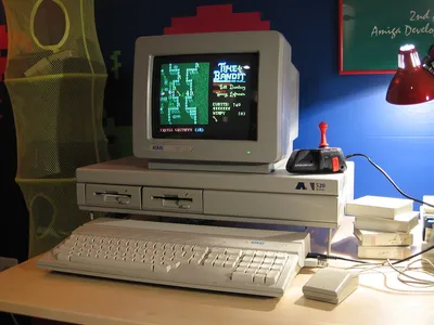 Vintage Atari 520ST computer setup displaying Time Bandit game on CRT monitor, with keyboard, joystick with red button, and mouse on a desk. Yellow speaker visible on left and red desk lamp on right in background.