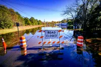 A flooded roadway leading to a 'Road Closed' sign flanked by orange traffic barrels, with floodwater stretching to submerged buildings and parked cars in the distance under a clear blue sky.