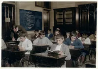 Colourized archival photo of a classroom full of boys at wooden desks, most bent over open books or writing with pens, in an early 20th-century schoolroom with a world map on the chalkboard and a teacher leaning over a student's desk in the background.