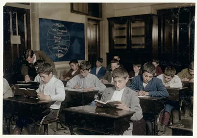Colourized archival photo of a classroom full of boys at wooden desks, most bent over open books or writing with pens, in an early 20th-century schoolroom with a world map on the chalkboard and a teacher leaning over a student's desk in the background.