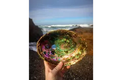 A hand holding an abalone shell up toward the camera on a rocky beach, displaying its iridescent interior of swirling greens, purples, pinks, and golds with characteristic respiratory holes visible on the left side. Crashing ocean waves and dark coastal rocks are visible in the blurred background under a partly cloudy sky.