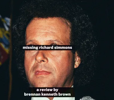 Extreme close-up crop of a colour photograph of Richard Simmons' face, showing his voluminous dark curly hair, forehead, nose, and mouth in warm, slightly grainy tones with a dark background. A colourful blurred flag is faintly visible in the upper left corner. Two black text bars overlay the image: one across the eyes reading 'missing richard simmons,' and one near the bottom reading 'a review by brennan kenneth brown,' both in bold white sans-serif type.