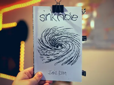Hand holding a poetry chapbook titled “sinkhole,” the cover clipped at the top with a black binder clip. The light blue cover shows a loose black-ink drawing of a swirling vortex or whirlpool, with the title written in rough, hand-lettered lowercase at the top and “Jake Beka” handwritten at the bottom. Patterned tape tabs stick out from the right edge of the booklet. The background is softly blurred with warm yellow string lights forming curved lines.
