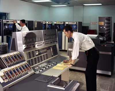 Two men in white dress shirts operate an IBM 7090 mainframe computer system in a large data centre. One man leans over the central operator's console writing on yellow notepad paper, while the other stands at a row of magnetic tape drive units in the background. The room is filled with large grey IBM mainframe cabinets and reel-to-reel tape drives. A numbered patch panel or channel selector is visible in the foreground left.