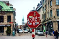 Un panneau d'arrêt rouge couvert de collants est planté au premier plan d'une rue pavée sur la place Jacques-Cartier, dans le Vieux-Montréal. Des deux bords, y'a des vieux bâtiments en pierre avec des toits mansardés pis des lucarnes, pis on aperçoit un clocher d'église au dôme vert en arrière-plan. La rue est pleine de chars stationnés pis d'autres en circulation, pis y'a quelques piétons qui marchent sur les trottoirs, près de boutiques comme la pizzeria « Bella Vita » pis le magasin « Jacques Cartier Souvenirs ». Le ciel est tout blanc et couvert, pis y'a un petit sapin en pot près de la devanture de gauche.