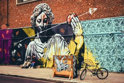 Une grande fresque murale sur un mur de briques représentant une figure de statue gréco-romaine classique rendue en noir, blanc et jaune, le regard baissé, tenant une pomme rouge à bout de bras. Des flèches et des livres ouverts flottent autour de la figure. Les sections de mur environnantes présentent des motifs décoratifs au pochoir : des fougères orange sur des fonds mauves et noirs à gauche, et de sombres motifs floraux et flocons de neige sur un fond bleu pâle à droite. Au premier plan, deux personnes sont assises sur le trottoir sous la fresque, une caisse en bois peinte d'un motif représentant une créature ailée et un véhicule se trouve à proximité, et une bicyclette est appuyée contre le mur. La fresque principale est signée « mateo » dans le coin inférieur gauche.