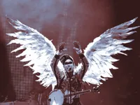 A musician performs on a dimly lit stage bathed in red and blue light, wearing an elaborate costume with large white-feathered angel wings spread wide behind them. They raise both hands overhead while singing into a microphone, wearing a patterned jacket and a banjo slung across their body. A black censor bar covers their eyes. Drum equipment is visible in the background.