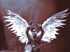A musician performs on a dimly lit stage bathed in red and blue light, wearing an elaborate costume with large white-feathered angel wings spread wide behind them. They raise both hands overhead while singing into a microphone, wearing a patterned jacket and a banjo slung across their body. A black censor bar covers their eyes. Drum equipment is visible in the background.