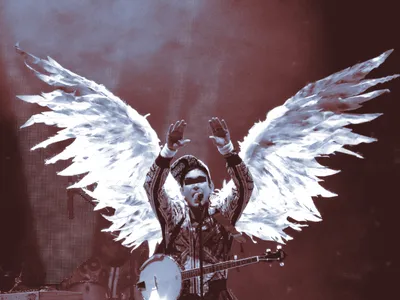 A musician performs on a dimly lit stage bathed in red and blue light, wearing an elaborate costume with large white-feathered angel wings spread wide behind them. They raise both hands overhead while singing into a microphone, wearing a patterned jacket and a banjo slung across their body. A black censor bar covers their eyes. Drum equipment is visible in the background.
