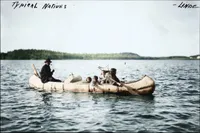 Colourized historical photograph of an Indigenous family traveling in a birchbark canoe on a calm lake. A man in a dark hat sits in the stern smoking a pipe, while a woman paddles at the other end. Three young children sit between them near a bundle of belongings, and one child reaches a hand toward the water. A forested shoreline lies in the distance under a pale sky. Handwritten at the top of the photo: ‘Typical Natives’ and ‘Linde’.