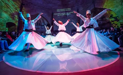 Whirling dervishes performing a traditional Sufi sema ceremony in a historic stone venue with arched architecture. Five performers in white robes and tall brown hats spin with arms outstretched, their wide skirts creating flowing circular patterns.