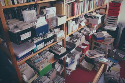 Wooden bookshelves overflowing with loose papers, documents, folders, and cardboard boxes including a Gabor shoe box. Additional stacks of papers and boxes are piled on a small side table and the red-carpeted floor; a knowledgeable home office or archive space.